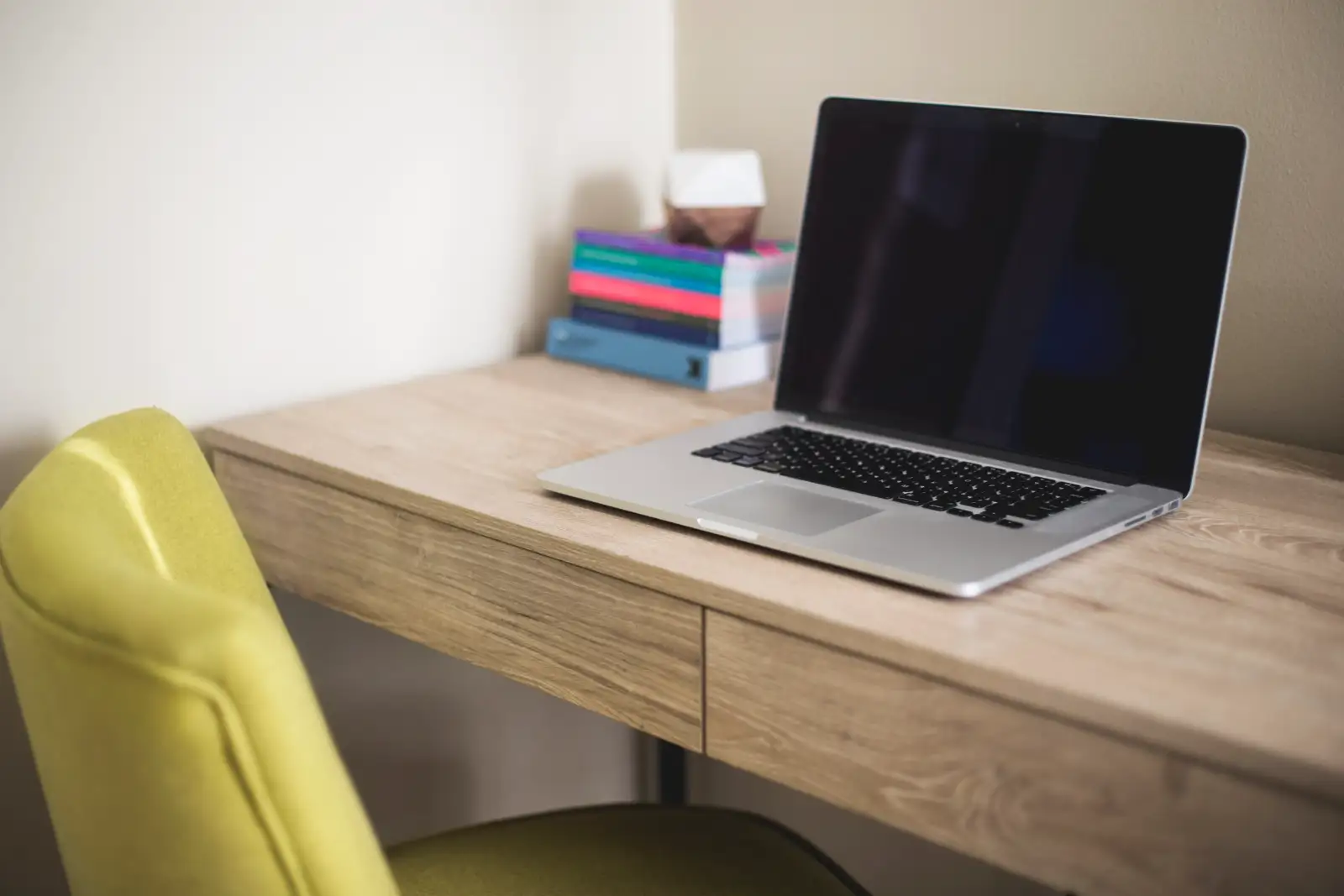 A laptop and a stack of books on a wooden desk beside a yellow chair